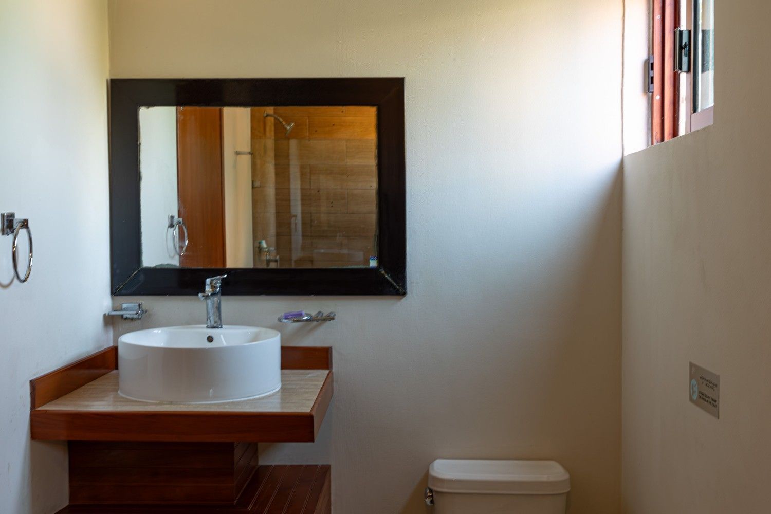Baño pequeño con lavabo blanco, espejo con marco oscuro e inodoro, con paredes de color beige.