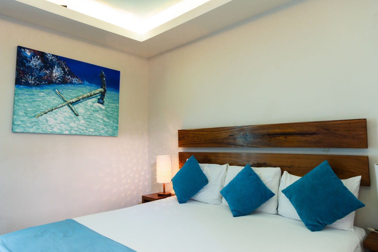 Dormitorio moderno con cama blanca, almohadas azules, cabecero de madera y cuadro de playa en la pared.