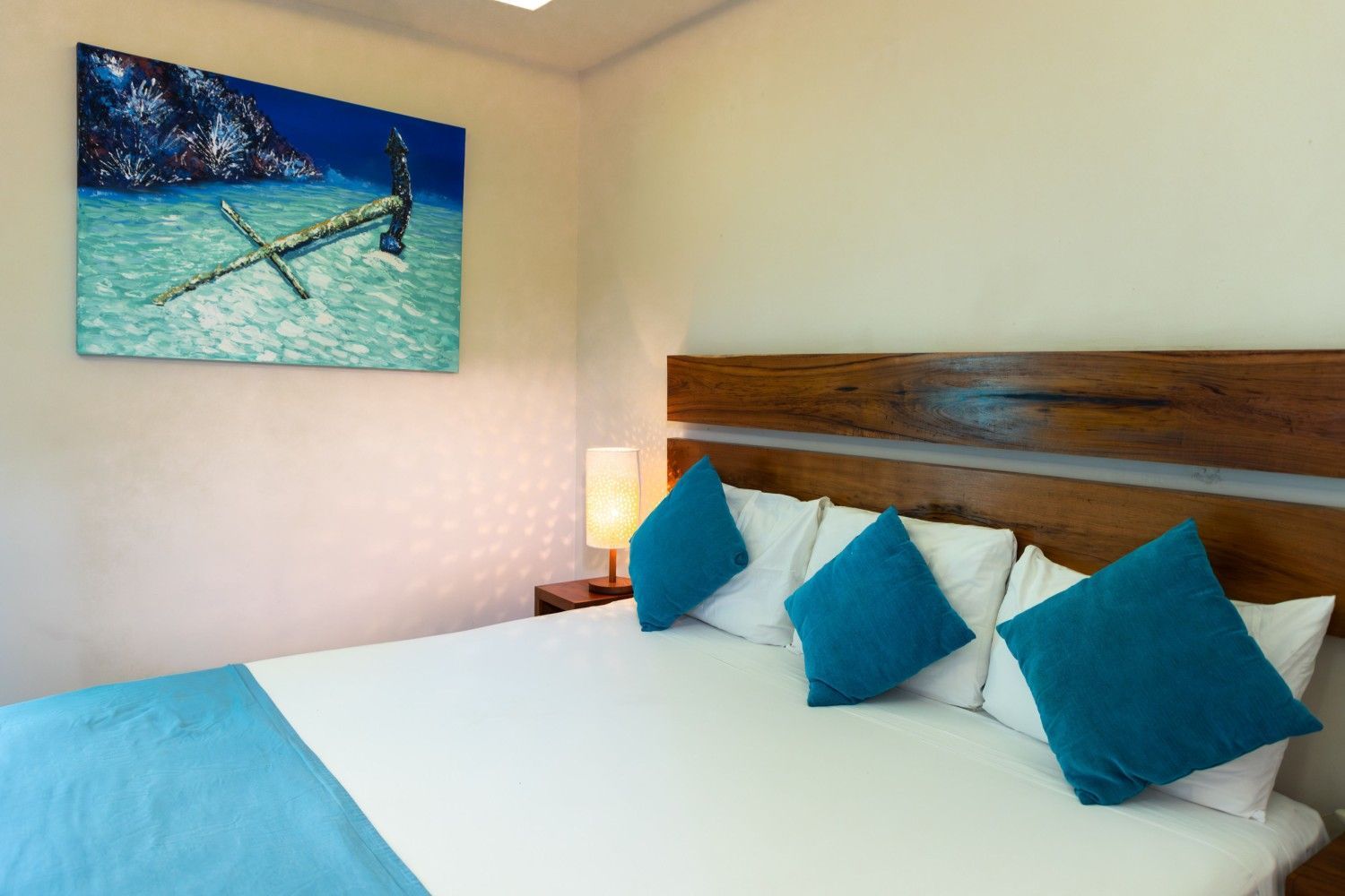 Habitación de hotel luminosa con cama blanca, almohadas azules, cabecero de madera y cuadro del océano en la pared.