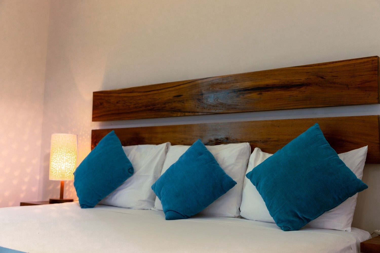 Cama de hotel con sábanas blancas, almohadas turquesas, cabecero de madera y lámpara de noche encendida.