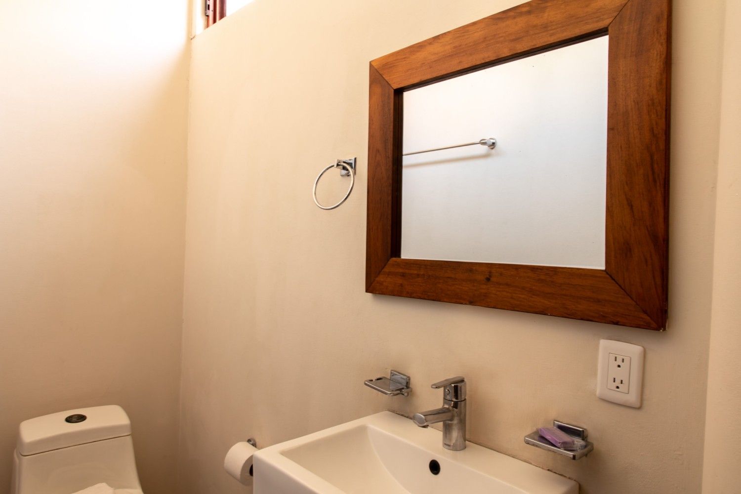 Baño pequeño con lavabo, espejo con marco de madera, inodoro, toallero y paredes de color beige.