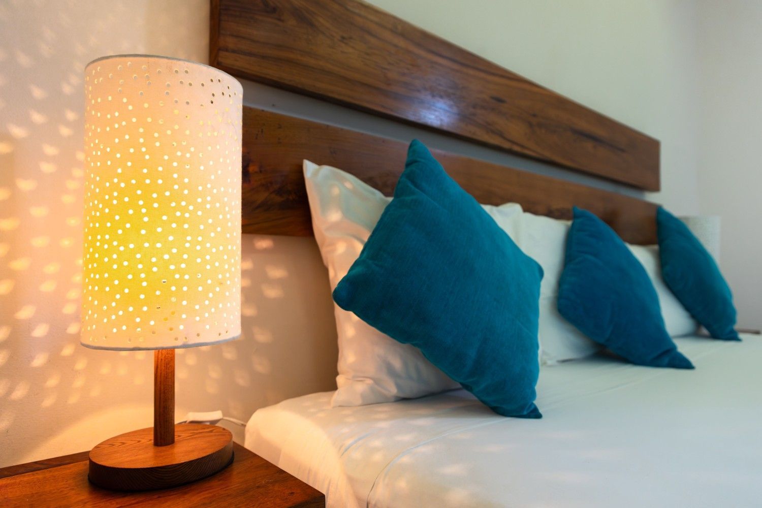 Cama con almohadas azules junto a una lámpara de noche con puntos luminosos sobre una mesita de noche de madera.