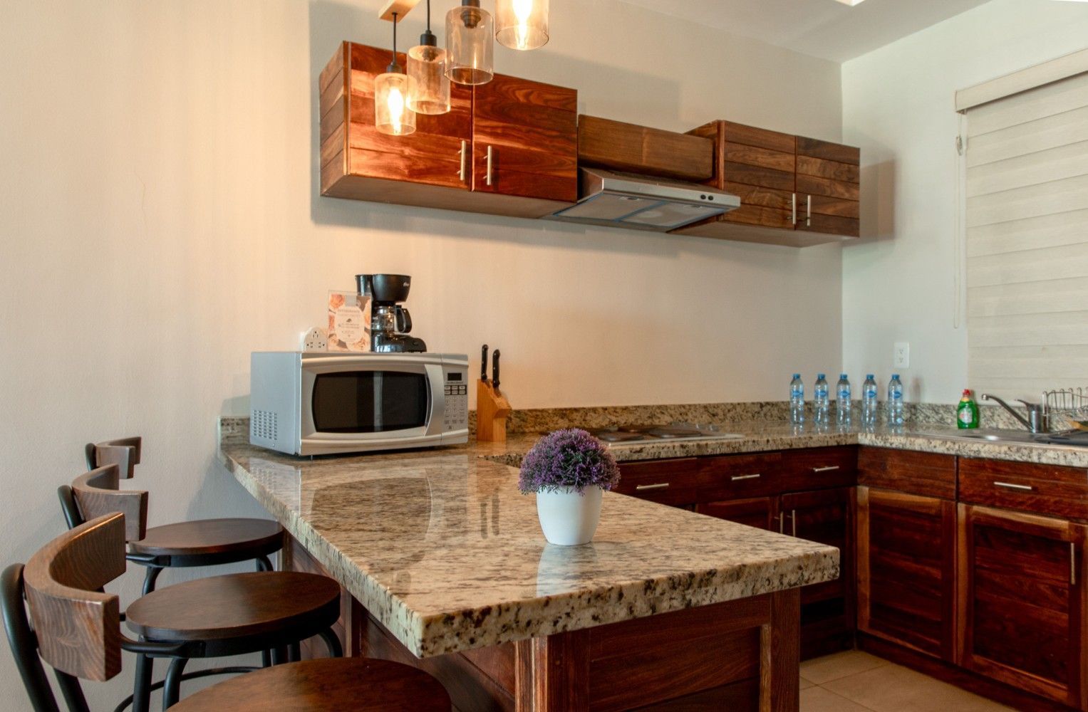Cocina moderna con isla de granito, gabinetes de madera, lámparas colgantes, microondas y taburetes de bar.