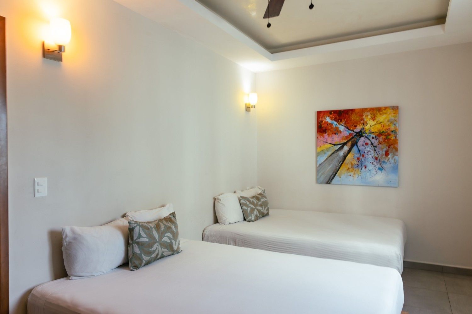 Habitación luminosa con dos camas individuales, paredes blancas, apliques de pared y obras de arte abstractas y coloridas.