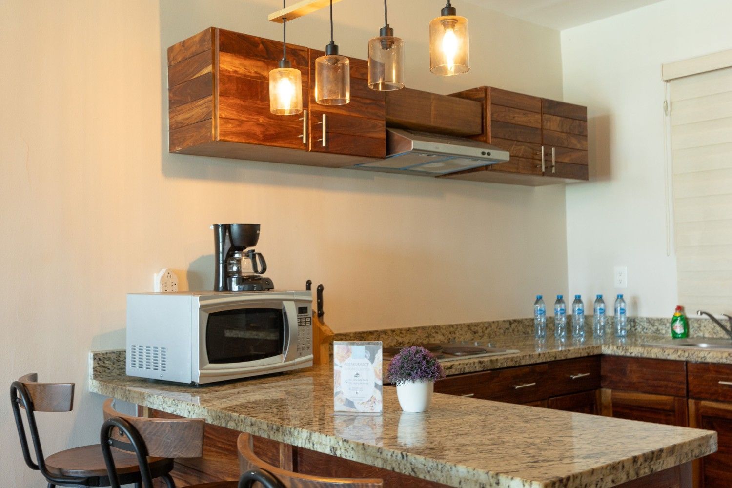 Cocina moderna con isla de granito, gabinetes de madera, lámparas colgantes, cafetera, microondas y botellas de agua.