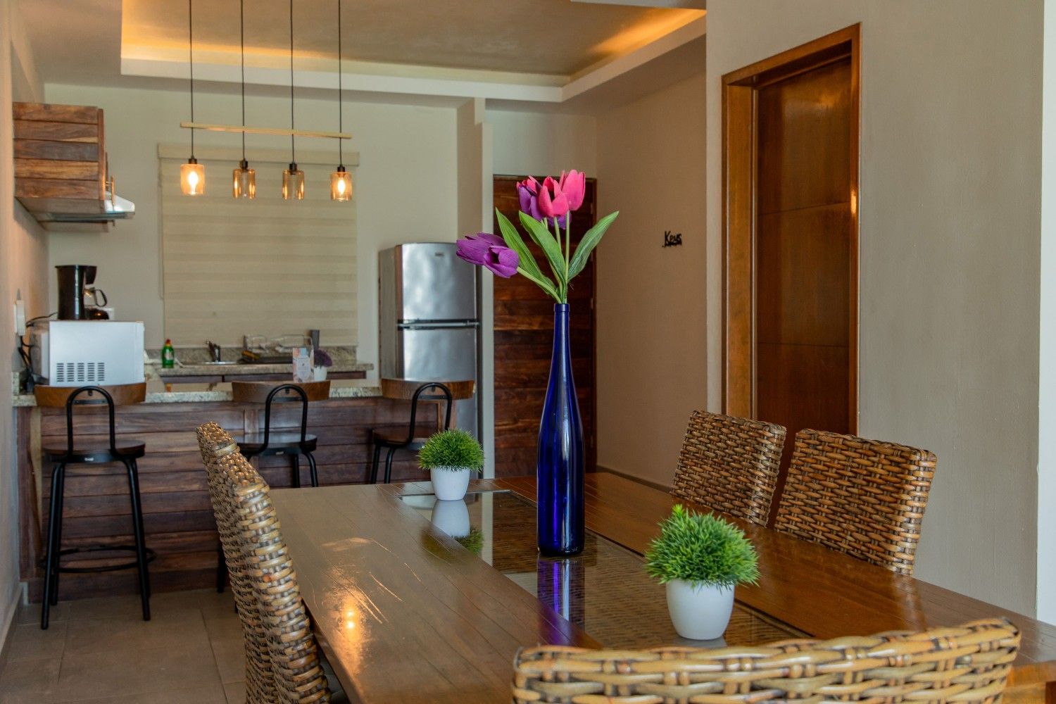 Comedor con mesa de cristal, sillas de mimbre y un jarrón con flores rosas en una cocina moderna.