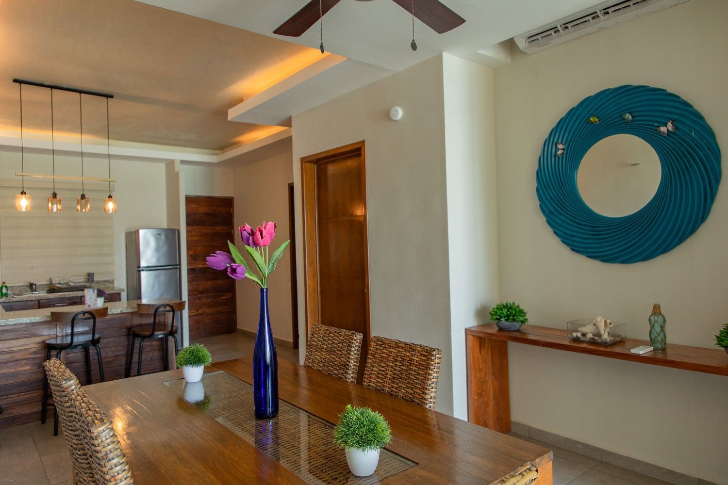 Comedor moderno con mesa de madera, lámparas colgantes, cuadro azul en la pared y jarrón con flores moradas.