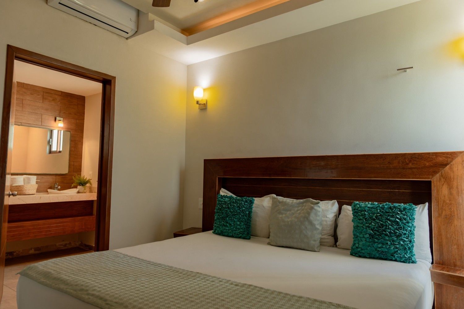Dormitorio con cama de madera, almohadas verdes, lámpara de pared, aire acondicionado y puerta abierta que da al baño.