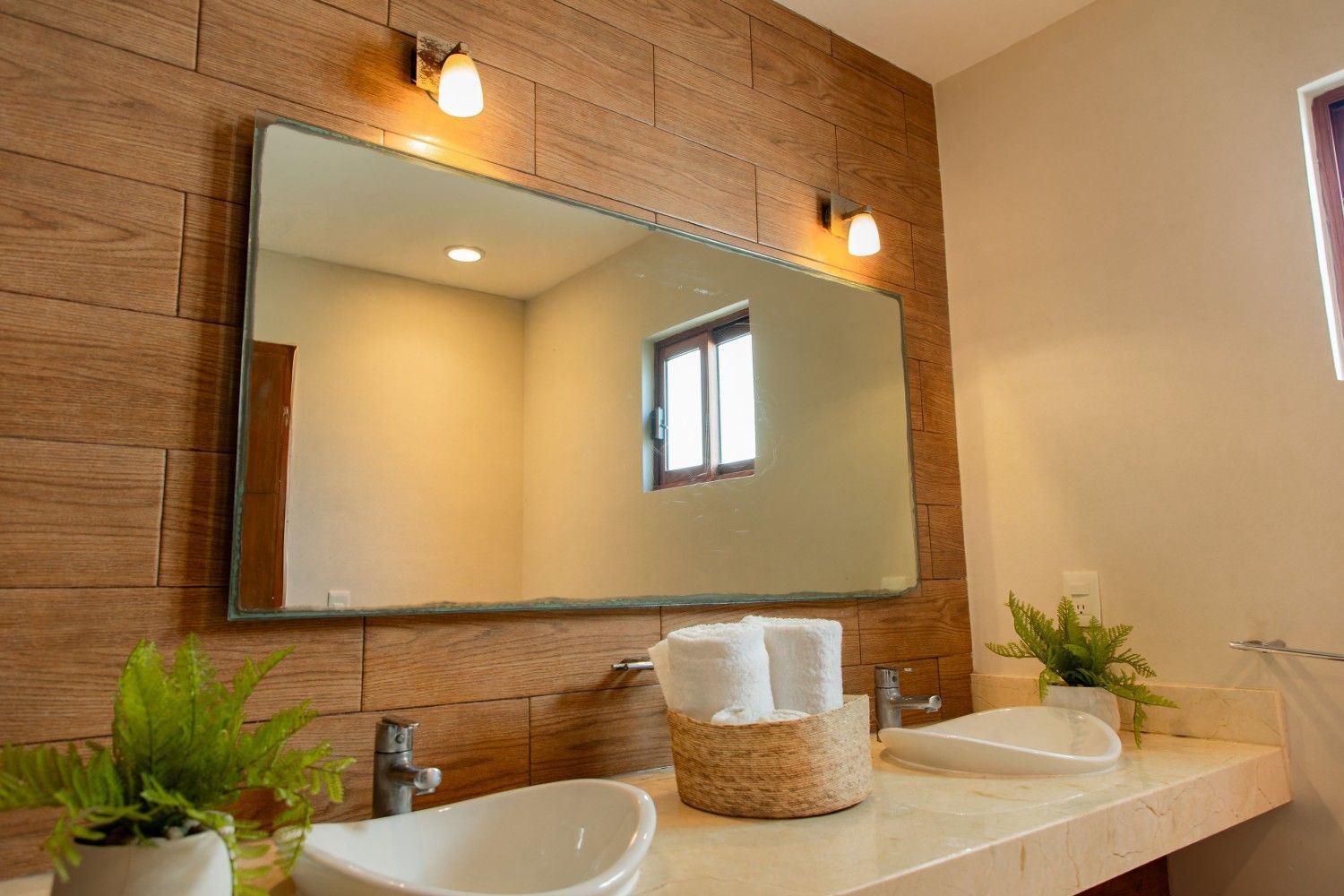 Tocador de baño moderno con espejo grande, apliques de pared, lavabo, plantas y encimera decorada con cestas tejidas.