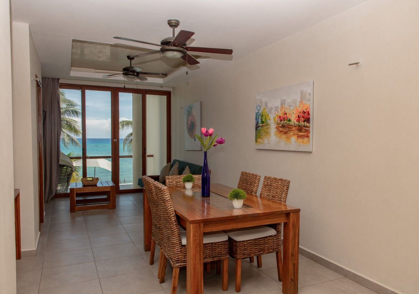 Comedor con mesa de madera, sillas de mimbre, ventilador de techo y balcón con vistas al mar.