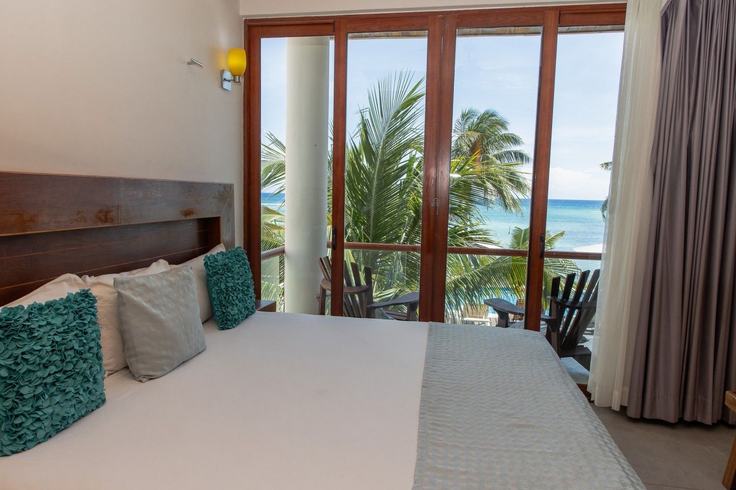 Habitación luminosa con cama king size, ventanas con marcos de madera y vistas al océano a través de las palmeras.
