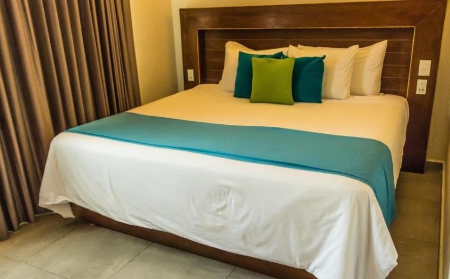Cama de hotel con sábanas blancas, camino turquesa y cojines verde azulados sobre cabecero de madera.