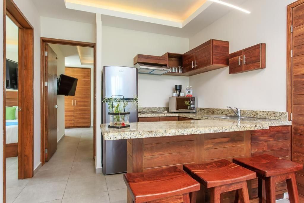Cocina moderna con isla de granito, taburetes de madera y nevera de acero inoxidable, vista desde un pasillo abierto.