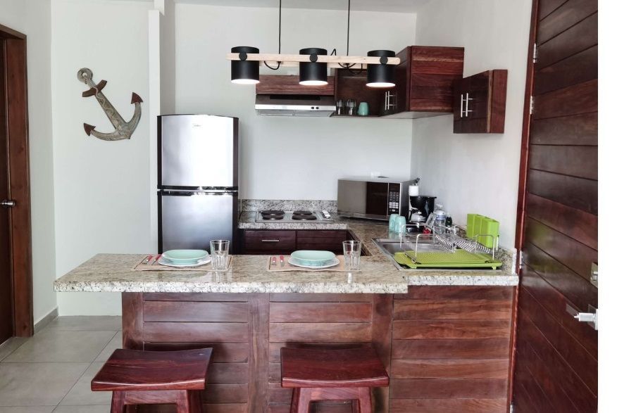 Rincón de cocina compacto con nevera de acero inoxidable, armarios de madera, encimera de granito y dos taburetes.