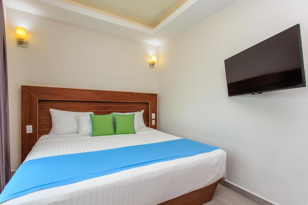 Habitación pequeña de hotel con cama de madera, colcha azul, almohadas verdes y televisor montado en la pared.