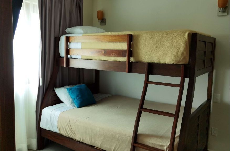 Litera de madera con ropa de cama beige y una almohada azul junto a una ventana con cortinas.