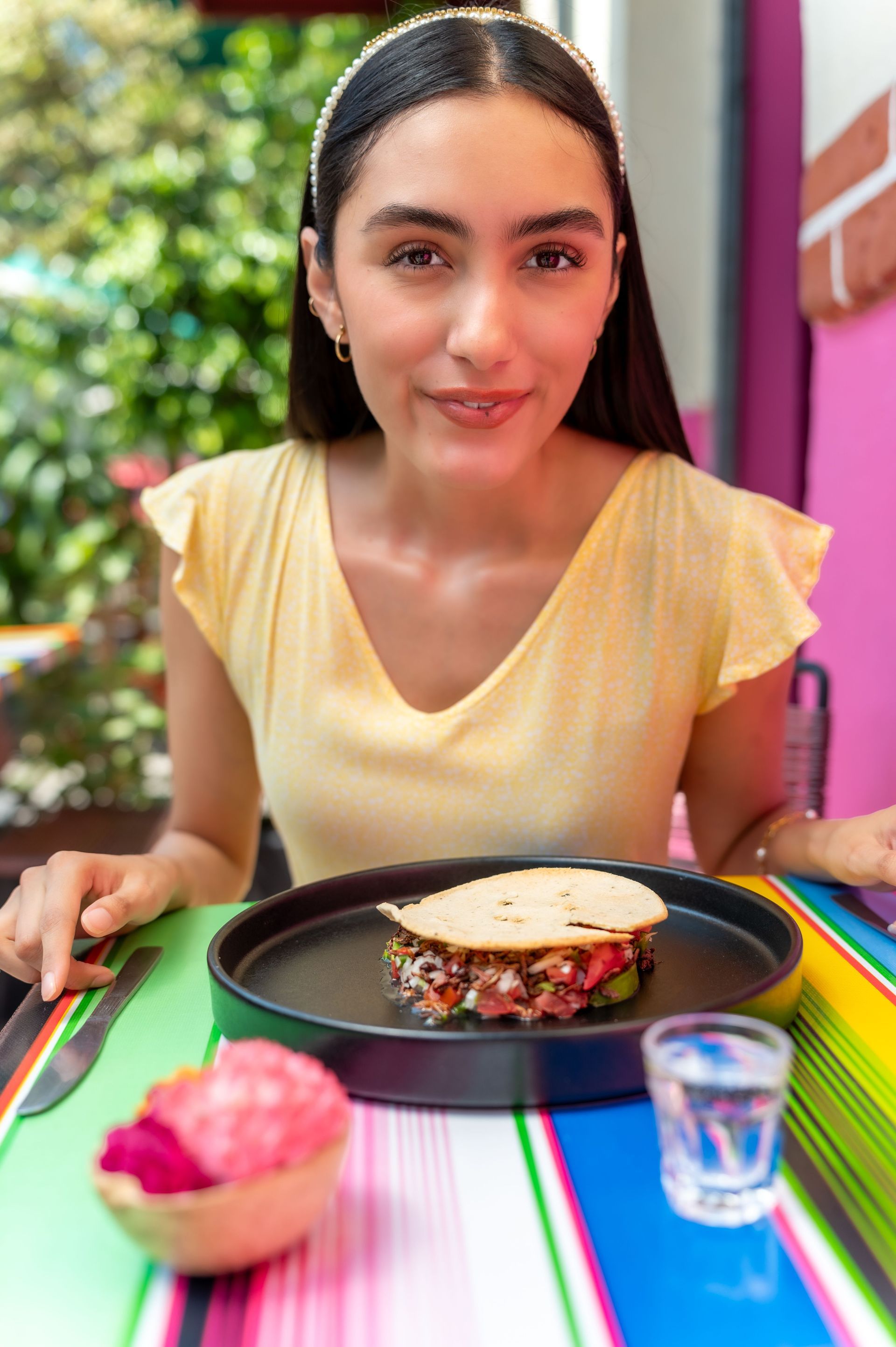 Mujer sentada en una mesa colorida con un plato de comida y una bebida pequeña al aire libre.