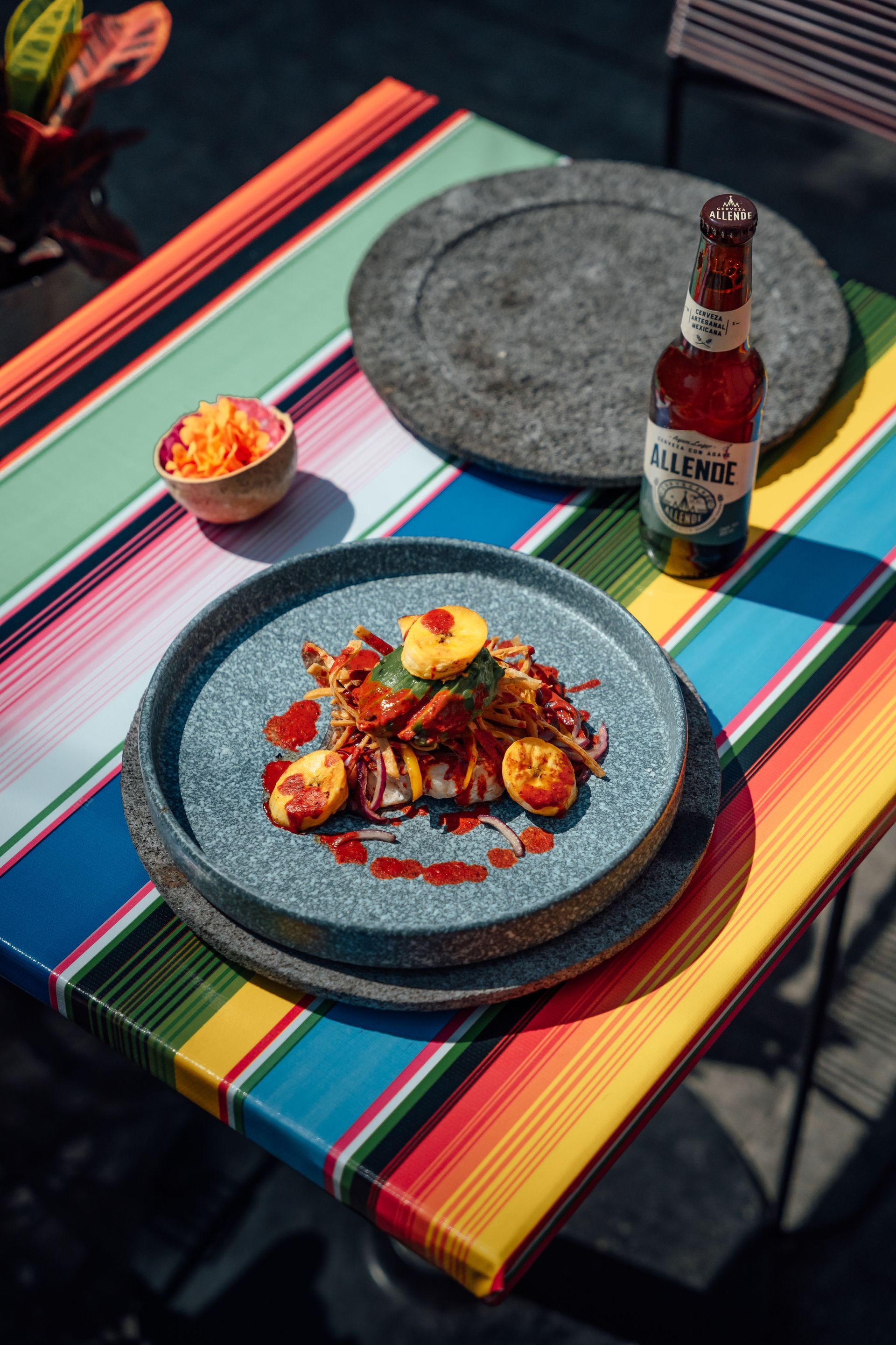 Dos platos de comida sobre un mantel de rayas de colores con una botella de salsa y un pequeño cuenco de guarnición.