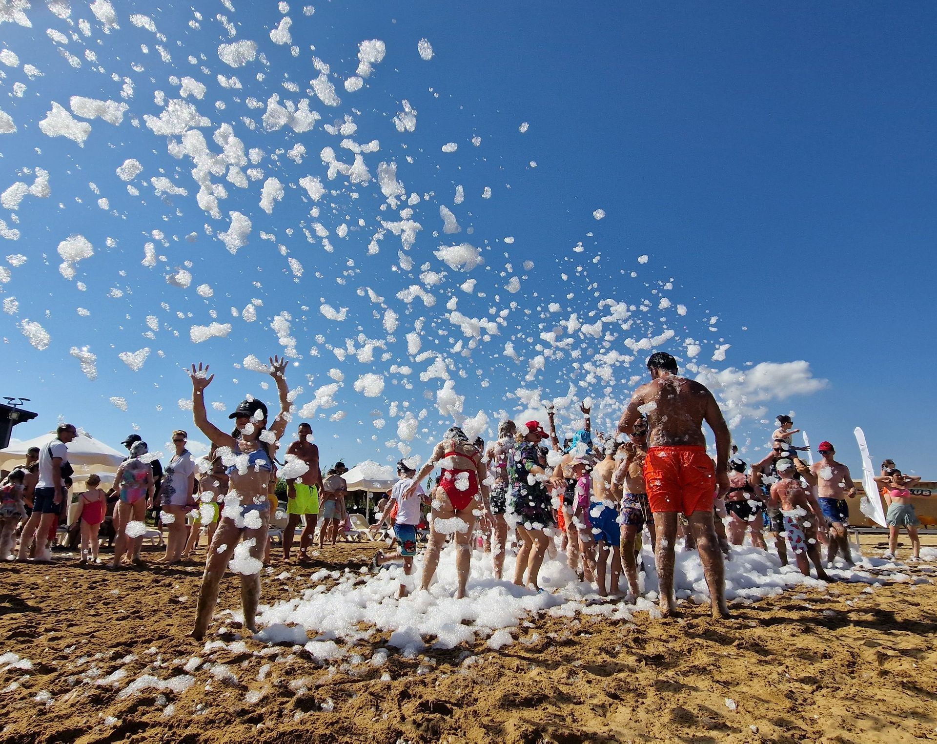 Multitud en una fiesta de espuma en la playa, gente vitoreando bajo un cielo azul despejado.