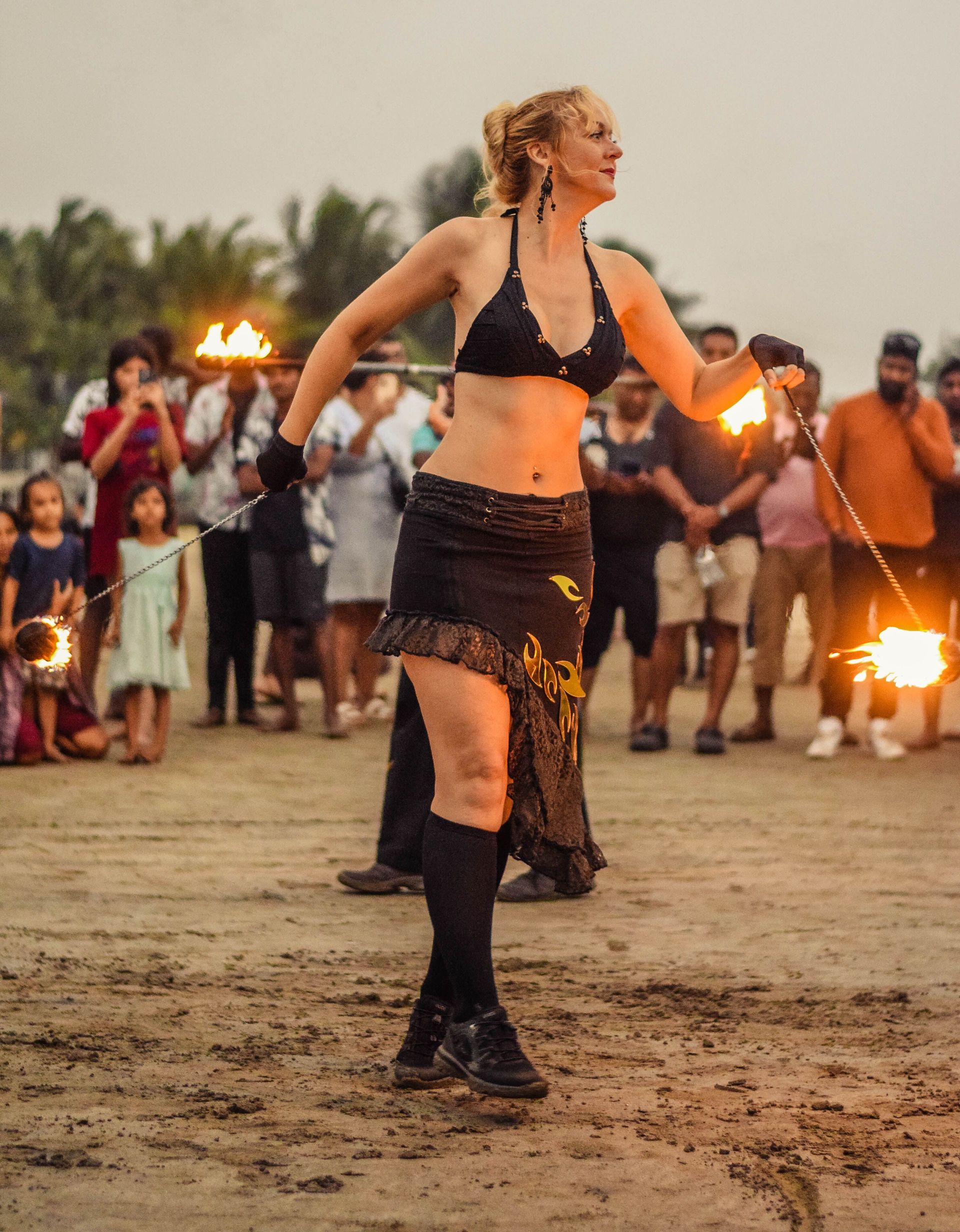 Bailarina de fuego actuando en una playa al atardecer, con llamas brillando al fondo.