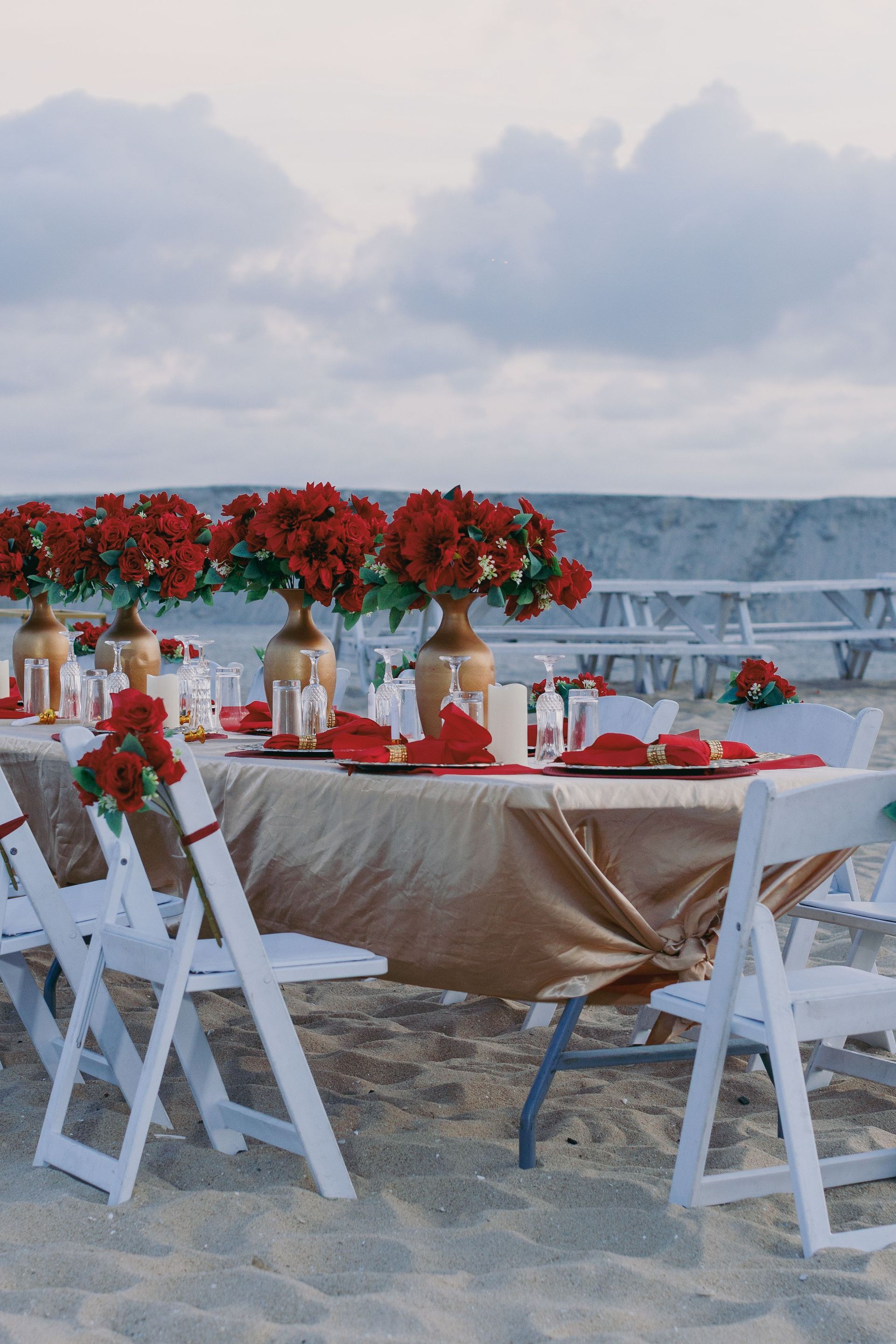 Mesa de comedor al aire libre con flores rojas y sillas blancas con vistas a la costa al atardecer.