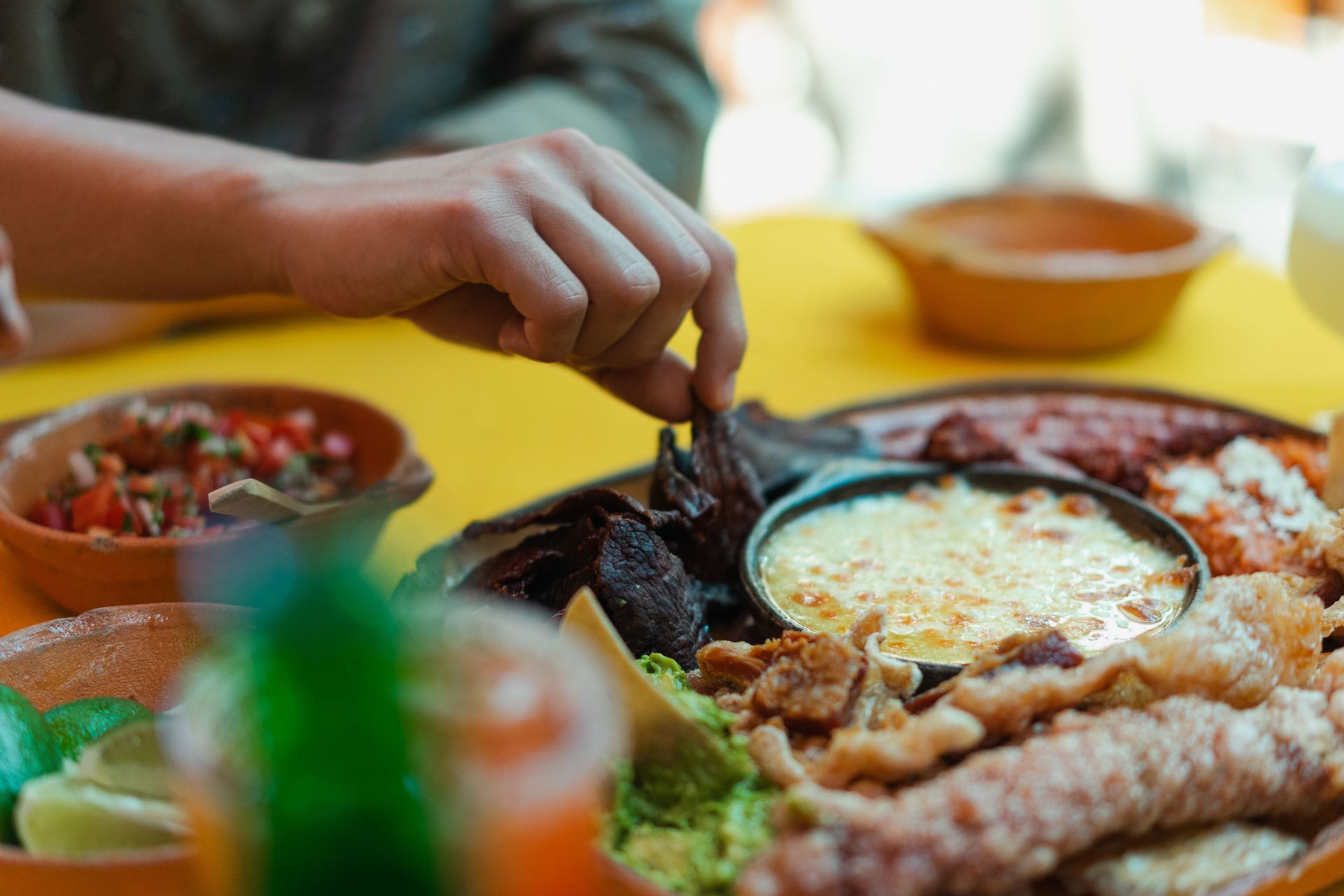 Una mano se extiende para alcanzar la comida en una mesa con un plato de queso al horno y guarniciones en un ambiente cálido.