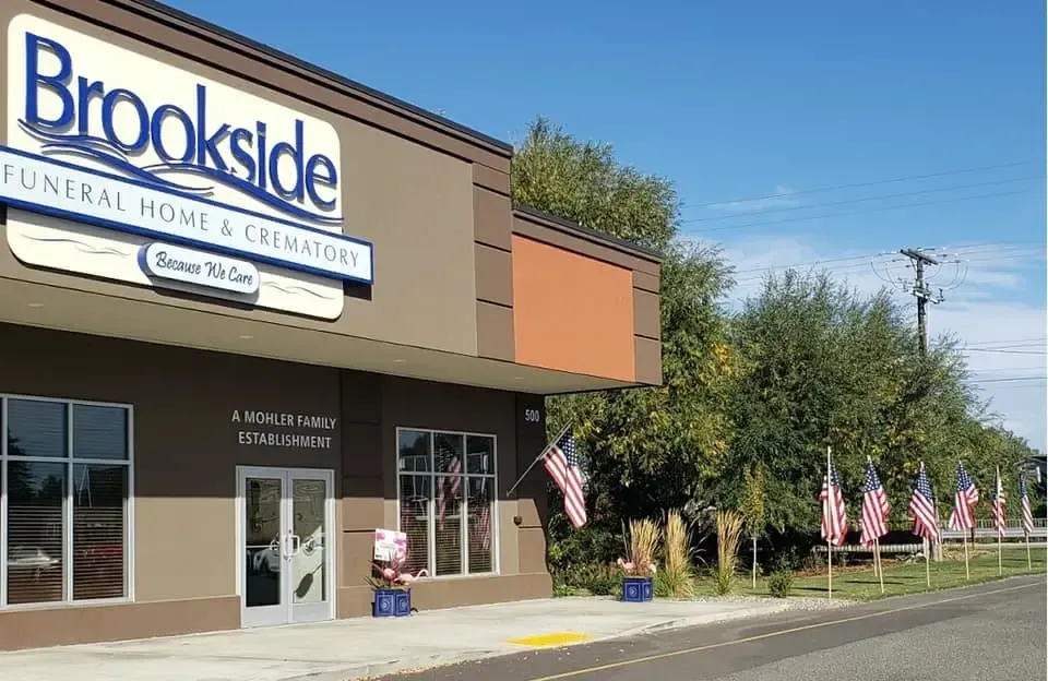 Brookside Funeral Home building with American flags. Brown and tan facade, blue sky, and trees.