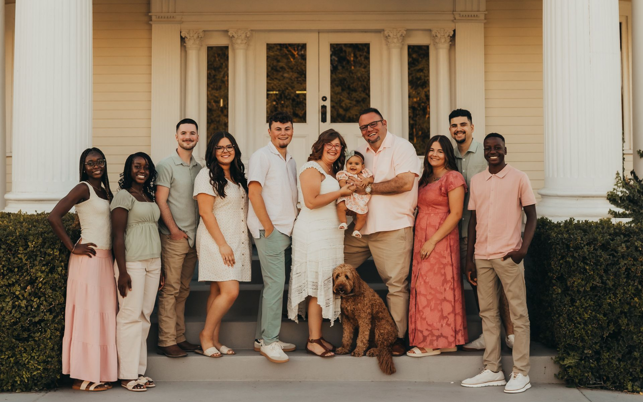 Group of people stand on porch of white house, posing for a photo.