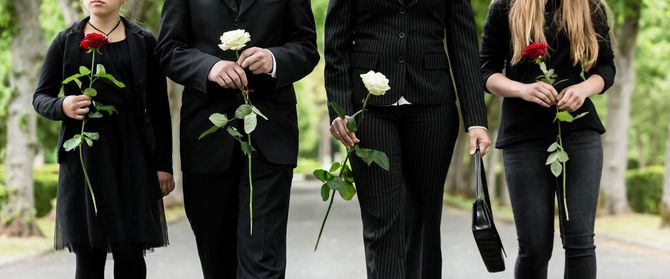 Four people in black clothing holding roses walk down a path lined with trees.