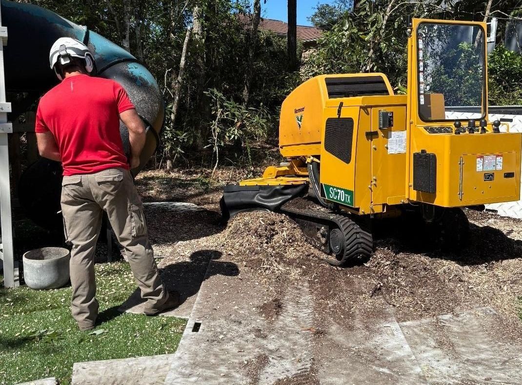 Man in red shirt operating a yellow stump grinder outdoors. Mulch surrounds the equipment.