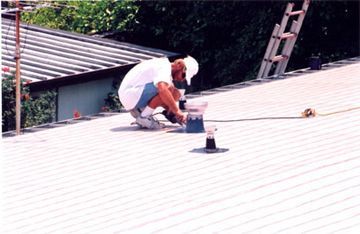 A man is kneeling on a roof with a ladder in the background