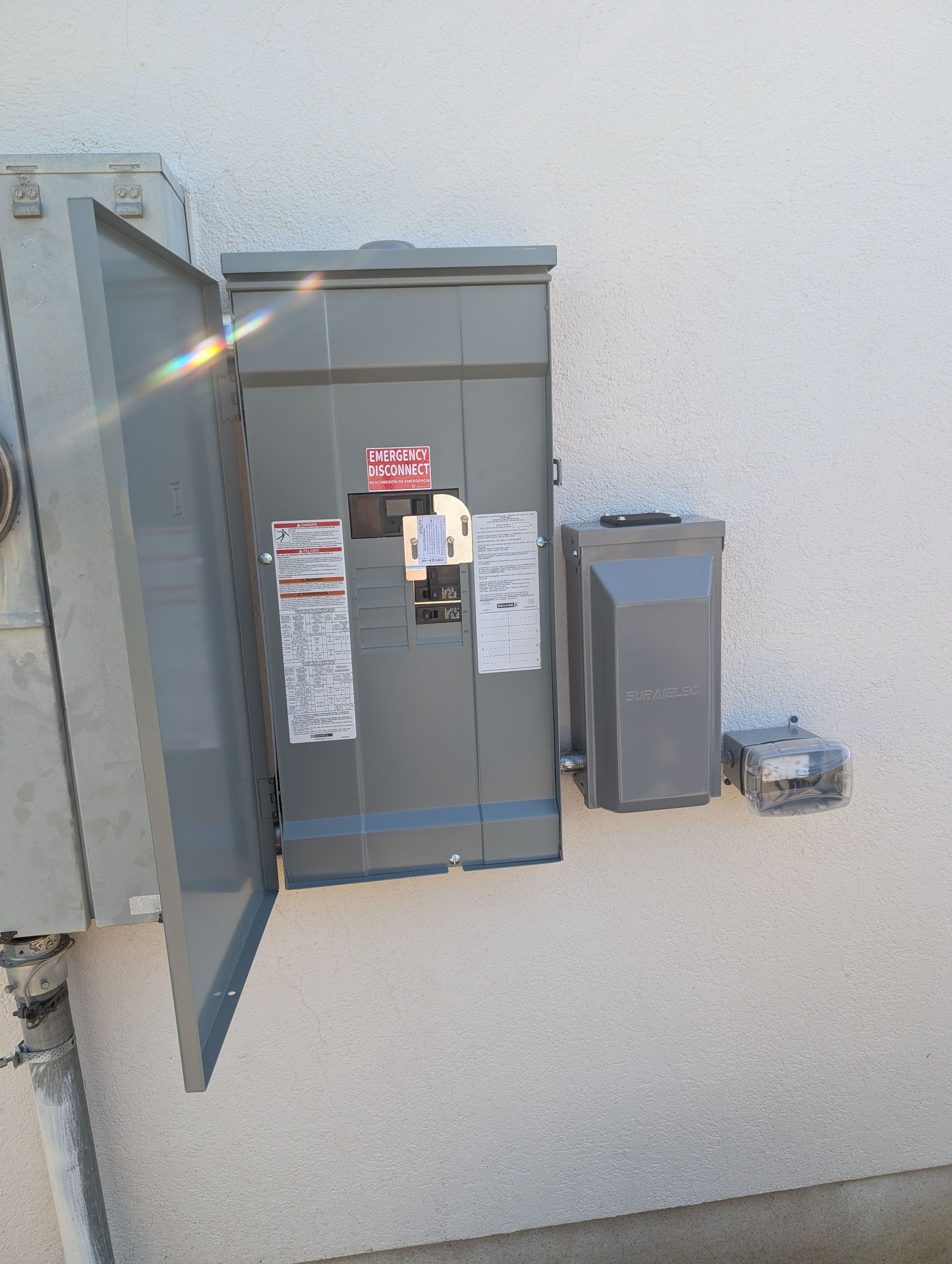Open electrical panels mounted on an exterior stucco wall with utility meters and boxes nearby