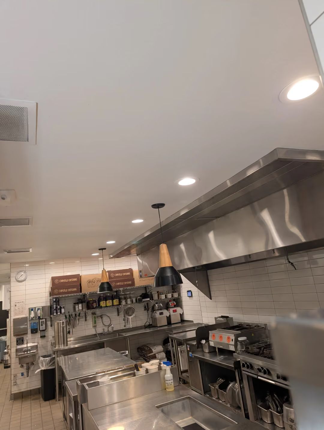 Commercial kitchen with stainless steel counters, ovens, and overhead hood under bright ceiling lights.