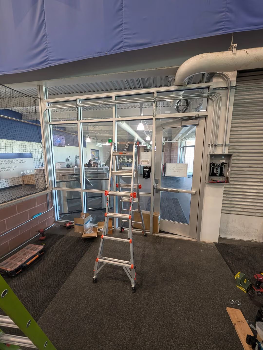 Ladder centered in a covered industrial walkway with scaffolding, pipes, and a glass entrance behind it.