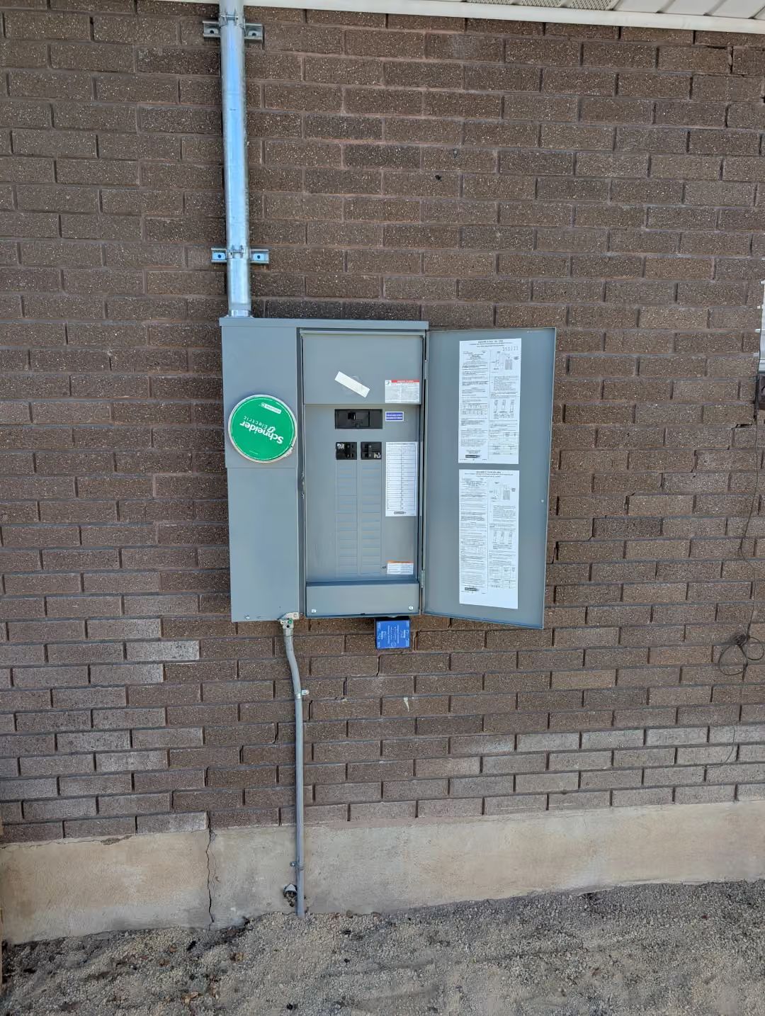 Gray utility electrical panel mounted on a brick wall with conduit and a green inspection sticker