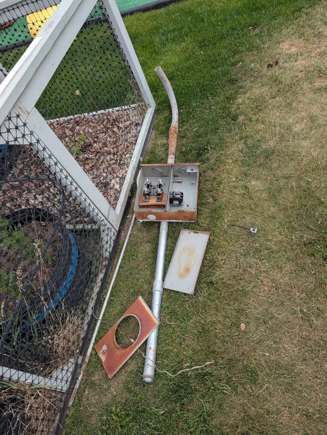 Broken electrical boxes and conduit on grass beside a chain-link fence.