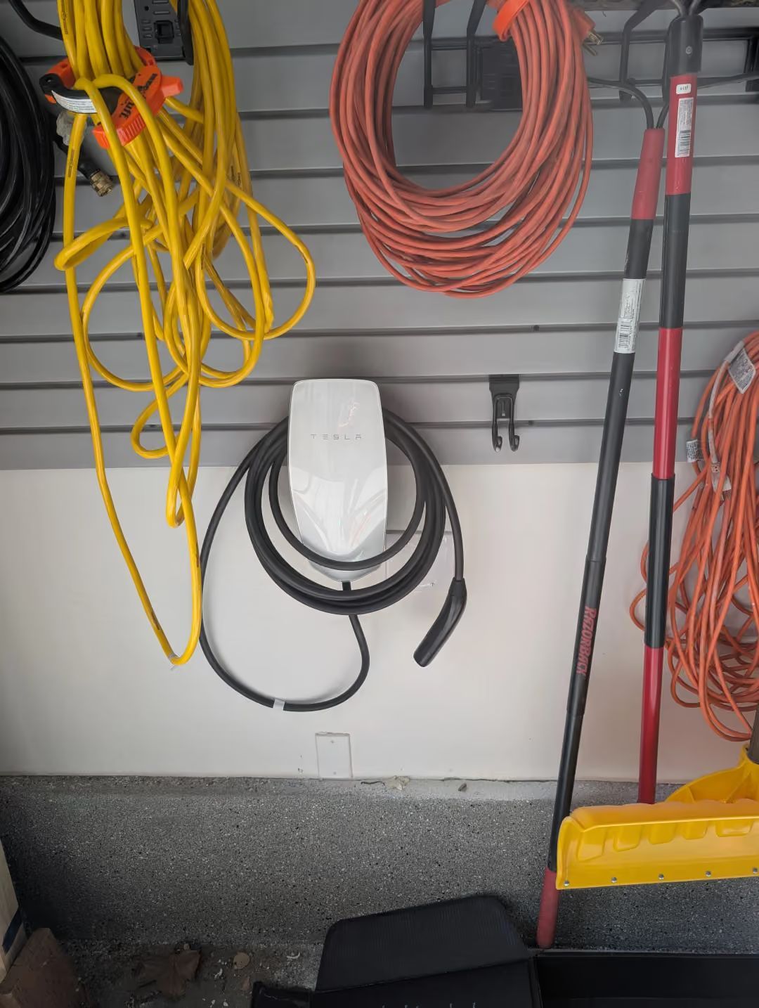 Garage wall with coiled cables, hanging cords, and tools on hooks above a concrete floor.