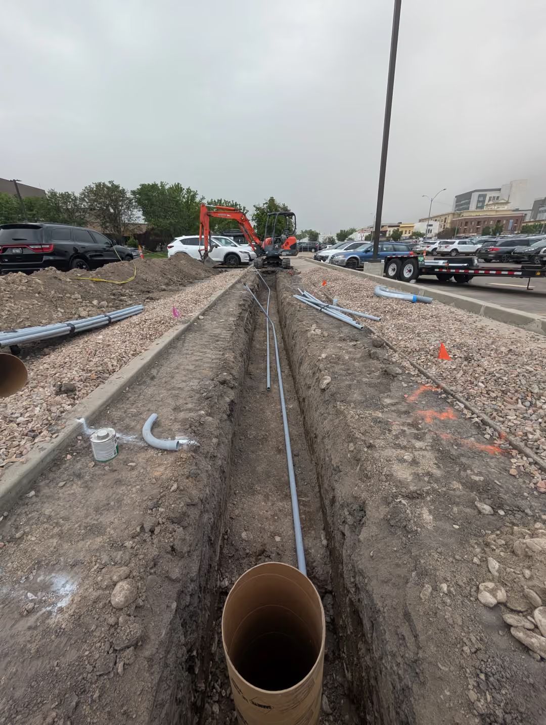 Trench in a construction site with exposed pipes and gravel, extending toward machinery in the distance