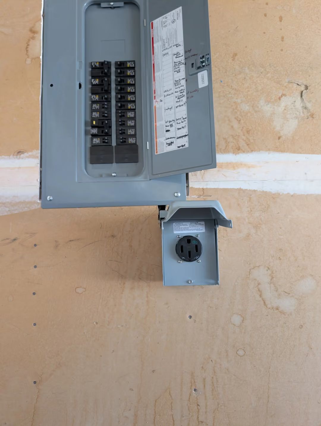 Open electrical breaker panel on a beige wall with a gray outdoor outlet below.