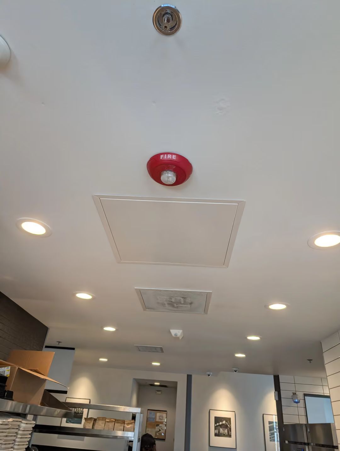 Ceiling view of a hallway with recessed lights, red fire alarm, and vents in a modern indoor space