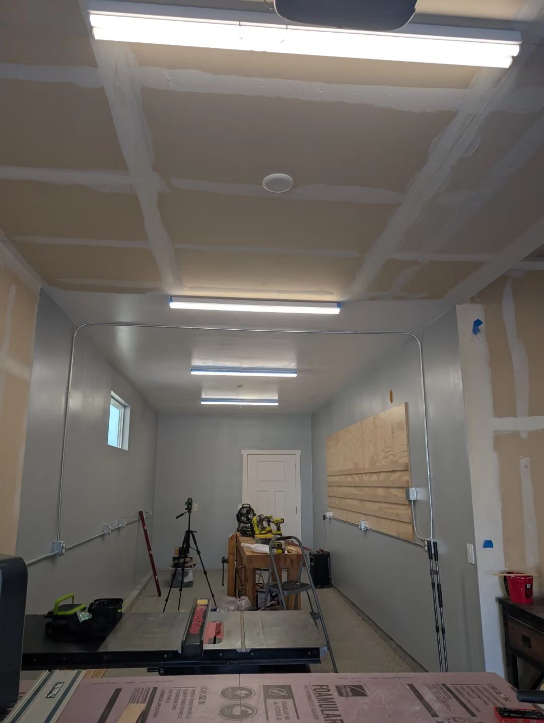 Narrow workroom with unfinished ceiling, fluorescent lights, tools, and a ladder against the right wall.
