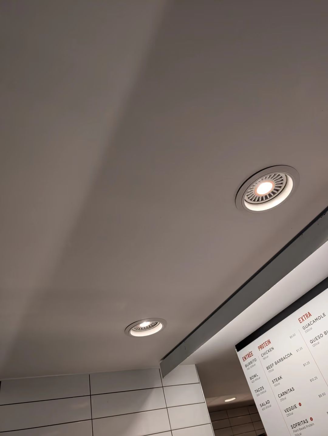 Ceiling with recessed lights above a white tiled wall and a partial menu board.