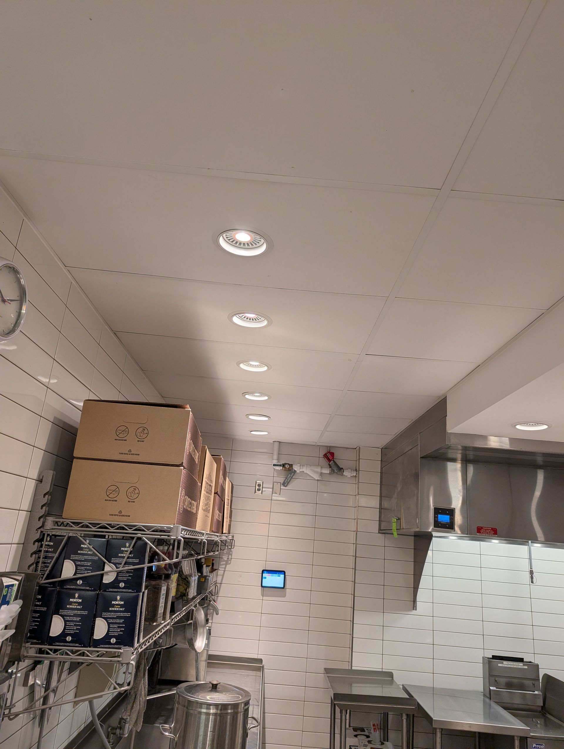 Narrow industrial kitchen aisle with shelves, boxes, and stainless steel appliances under recessed ceiling lights