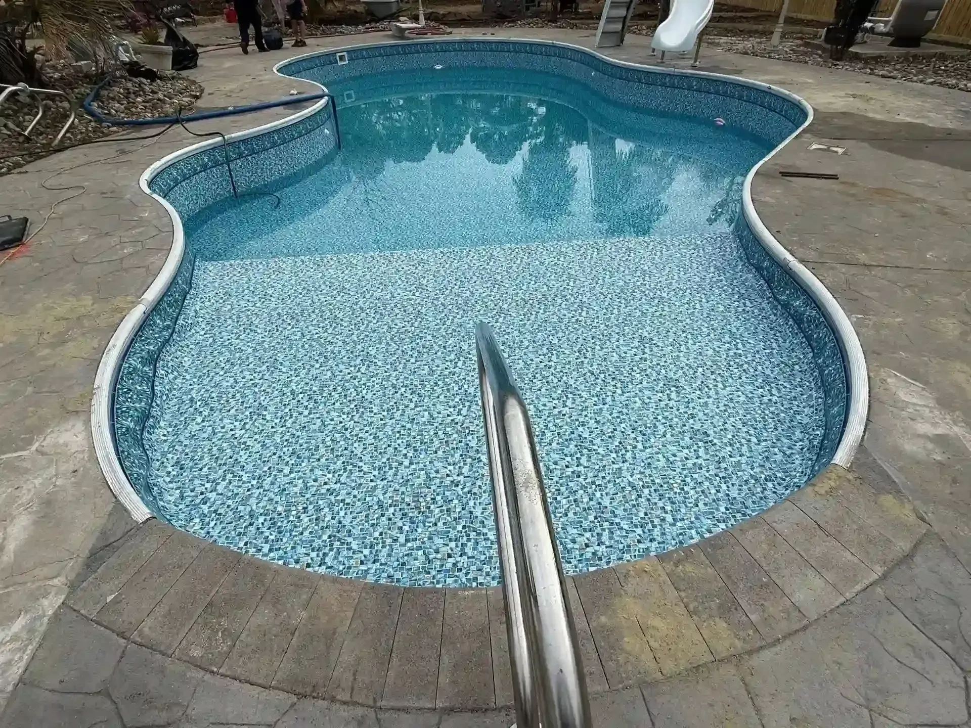 Curved backyard swimming pool with blue mosaic tile and stone deck, viewed from metal handrail area