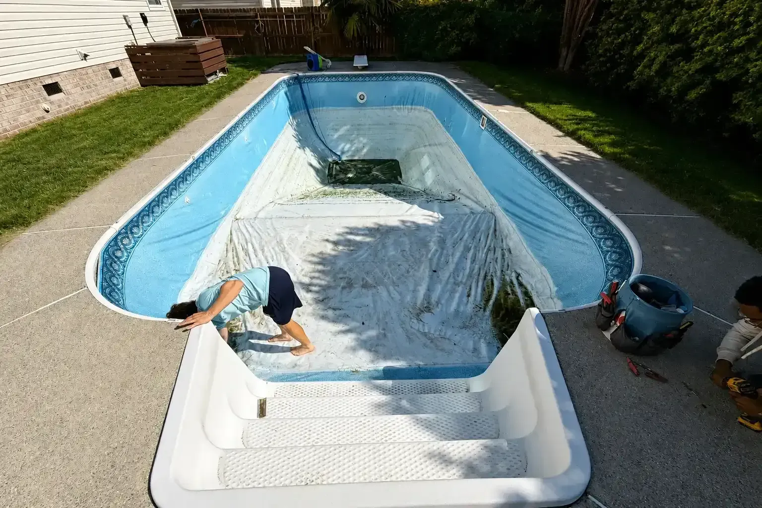 Backyard pool with white liner partially removed, showing workers at the edge near a house and lawn