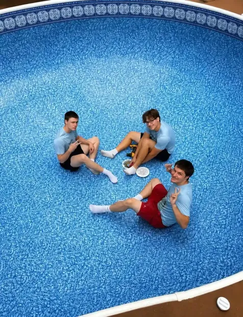 Three people sitting in an empty blue swimming pool, wearing light blue and red clothes.