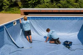 Two workers repair a drained blue swimming pool, one standing and one kneeling with tools.
