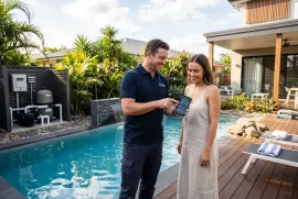 Man and woman by a pool, looking at a phone on a sunny patio with a modern house backdrop
