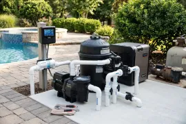 Pool pump and filtration equipment on a patio beside a swimming pool and garden shrubs.