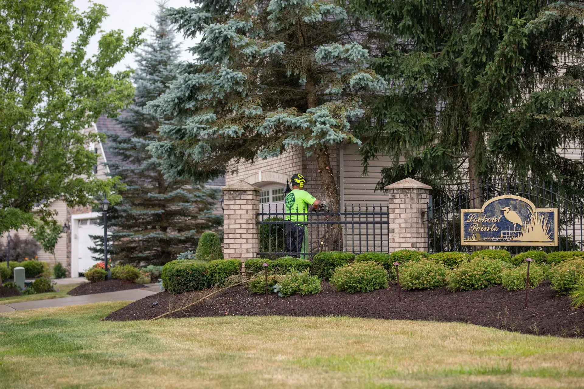 Landscaper trimming bushes at a gated community entrance with a sign, surrounded by landscaping.